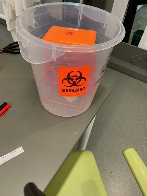 a benchtop wire container with hinged lid containing an orange biohazard bag inside a plastic secondary container