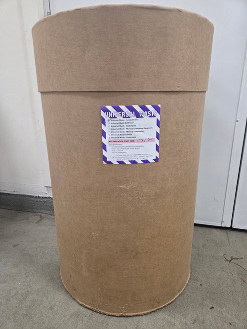 55-gallon brown fiberboard drum with lid and purple and white label.  Label is marked with Universal Waste Aerosols and Accumulation Start Date of 7/31/2024