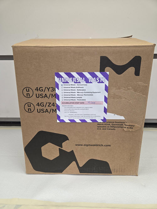 Brown fiberboard box with closed flaps and purple and white label.  Label is marked with Universal Waste Aerosols and Accumulation Start Date of 5/1/2025